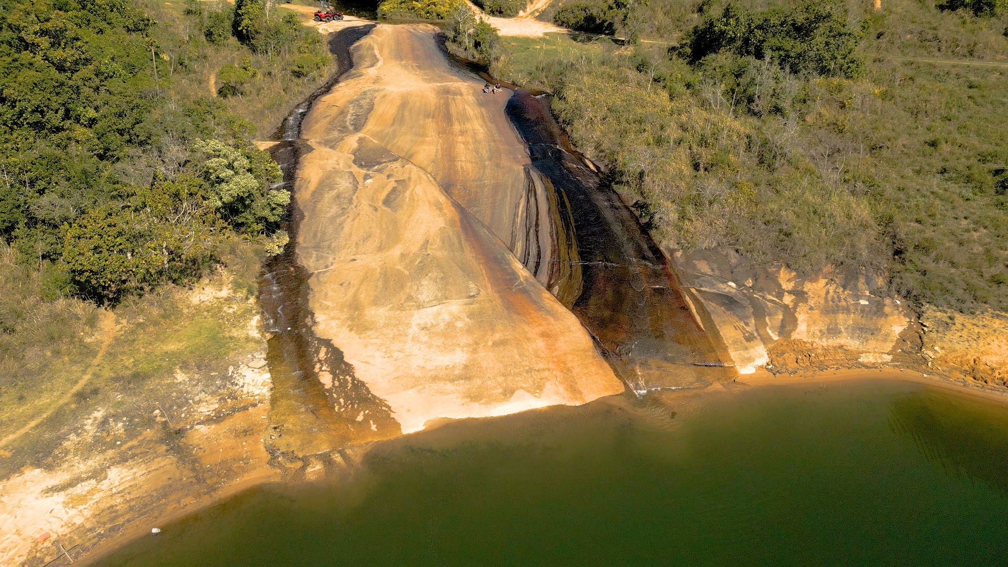 (Vista aérea da nascente do Rio Tietê em Salesópolis, São Paulo, mostrando a água esverdeada que emerge na base de uma formação rochosa inclinada e escorregadia. A imagem destaca o contraste entre as cores da pedra, com tons de marrom, preto e ocre, e a vegetação verdejante que circunda o local. Dois aventureiros são visíveis no topo da formação, perto de quadricíclos vermelhos, evidenciando o local como ponto turístico e destino para atividades ao ar livre durante a Aventura General Tire. Esta nascente é um marco natural importante, simbolizando o início do icônico Rio Tietê)