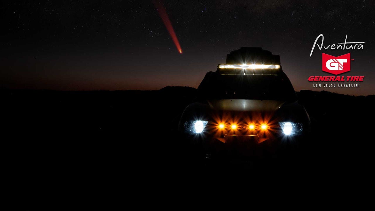 A imagem mostra um céu noturno estrelado, com um cometa brilhante cruzando o horizonte. No centro, há um veículo com os faróis acesos, destacando-se na escuridão. No canto direito, aparecem os logos "Aventura General Tire com Celso Cavallini". O Cometa é o C/2023 A3 (Tsuchinschan-ATLAS).