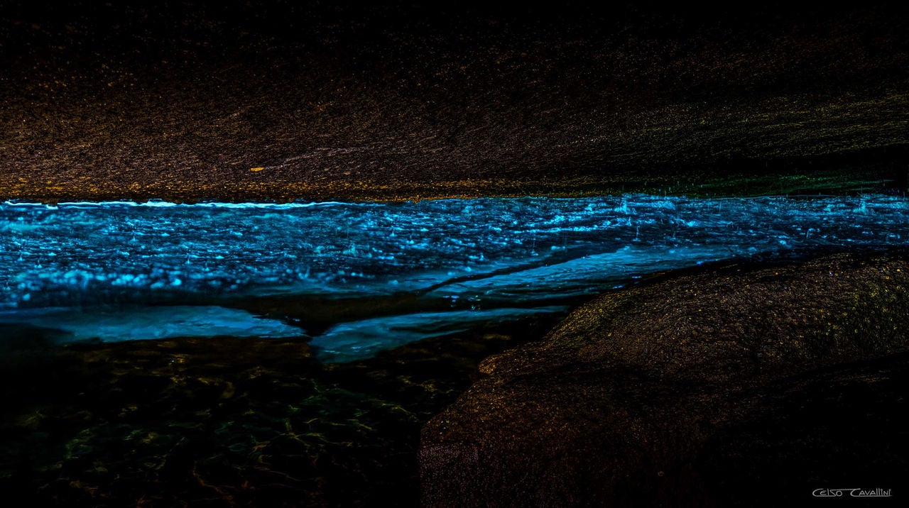 A imagem mostra o interior da Gruta Acaiá, com um reflexo de luz azul brilhante sobre a água escura, criando um efeito visual deslumbrante. A parte superior da caverna é escura e rugosa, enquanto a luz azul realça a superfície da água, transmitindo uma sensação de mistério e beleza natural.