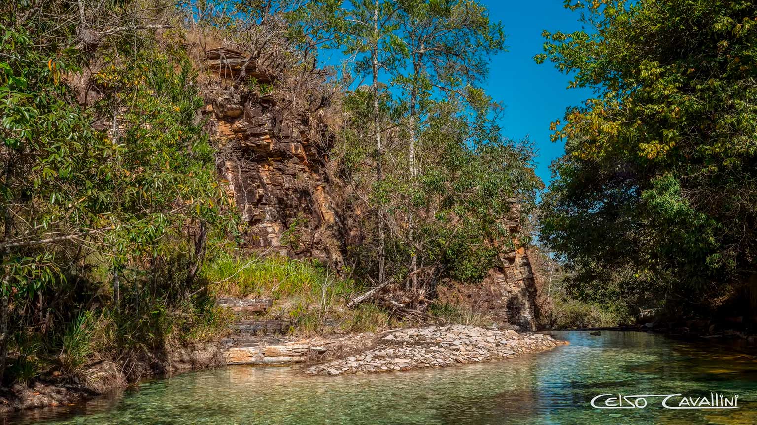 A imagem mostra um cânion cheio de água cristalina na coloração verde. Ao redor existe uma mata verde e o céu azul sem nuvens.