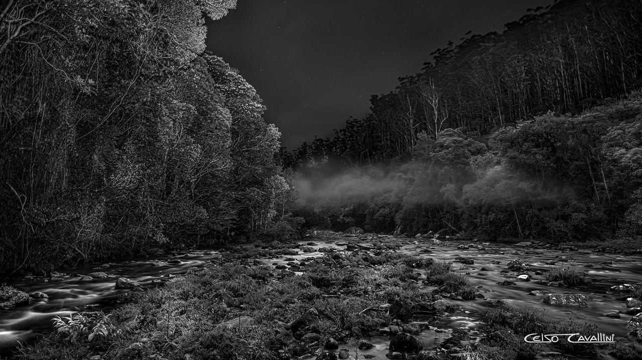 A imagem mostra um foto preto e branca tirada dentro do Parque das Neblinas. Na imagem é possível ver o Rio Itatinga e uma infinidade de vegetação ao redor do rio.