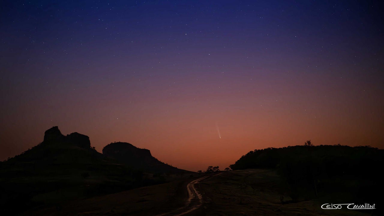 A imagem retrata uma paisagem noturna em uma atração turística chamada Três Pedras, em Bofete – município localizado no interior de São Paulo. Existe um céu estrelado e o cometa C/2023 A3 (Tsuchinschan-ATLAS) visível no horizonte, deixando um rastro luminoso no céu. O terreno é montanhoso, com duas grandes formações rochosas escuras em silhueta ao fundo. Um caminho de terra atravessa a imagem em direção ao horizonte. O céu apresenta uma transição de cores, indo do azul escuro no topo para tons alaranjados perto do horizonte, sugerindo o crepúsculo.