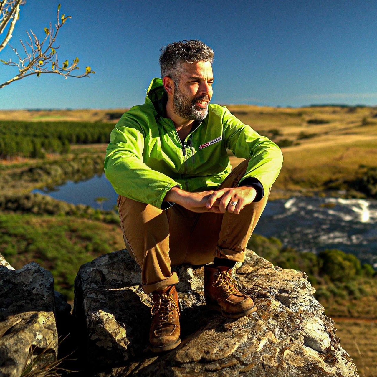 A imagem mostra o influenciador de expedições off-road Ricardo Pocholo sentado em uma pedra num mirante. O cenário atrás possui um rio e serras que vão até o horizonte.
